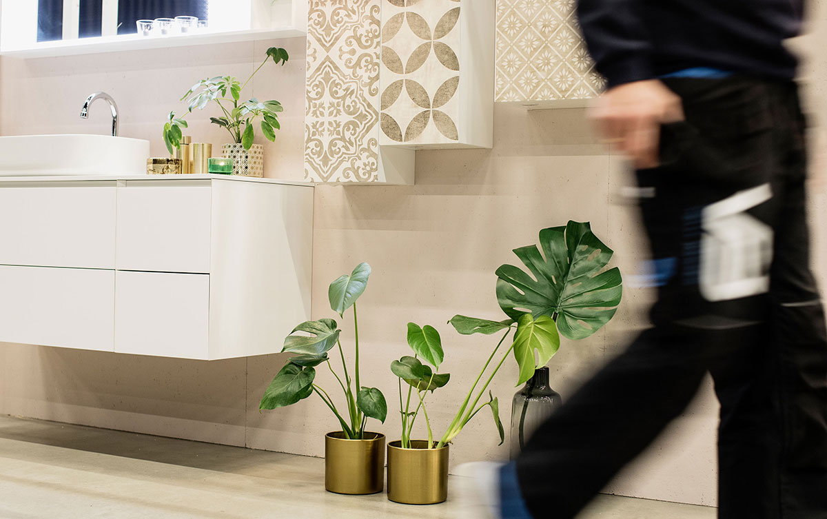 Baderom med servant og servantskap montert på beige flislagt vegg med skap i matchende mønster. Rørlegger i arbeidsantrekk i bevegelse ut av bildet.